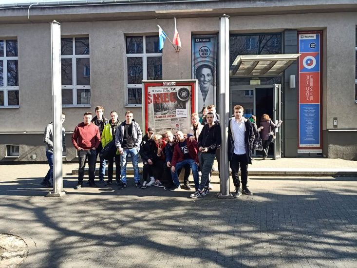 Grupa uczniów stoi przed budynkiem teatru Łaźnia Nowa. W tle wejście,  a za uczniami reklama.