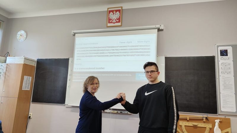 Sala szkolna. Na ekranie wyświetlone liczby. Obok ekranu stoją laureaci konkursu