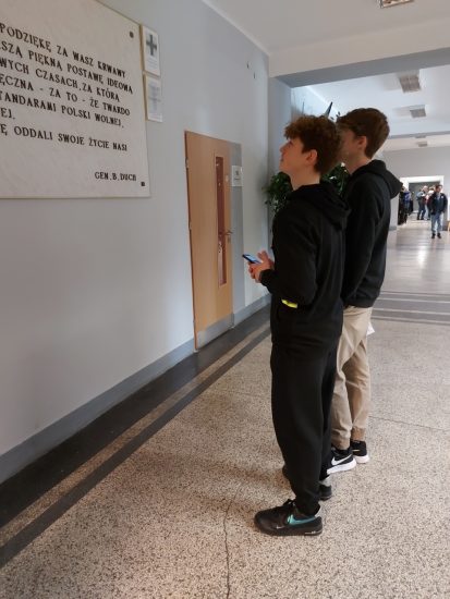 Dwóch uczniów stoi w holu szkoły przed tablicą ze słowami gen. Bronisława Ducha.