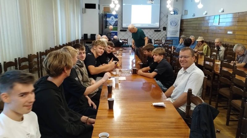 Sala wykładowa. Na ścianie wisi ekran. Uczestnicy seminarium siedzą przy dwóch stołach. 