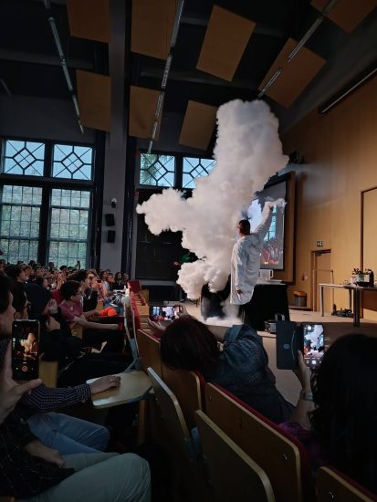 Sala wykładowa. Widać unoszącą się chmurę dymu (pary). Obok stoi naukowiec w białym fartuchu. 