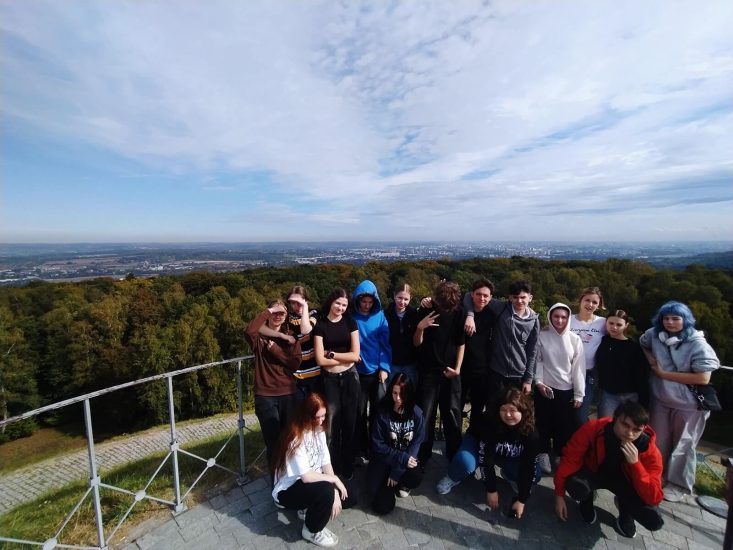 Grupa uczniów stoi na szczycie Kopca Kościuszki. Niektórzy opierają się o metalową barierkę. W tle panorama Krakowa.