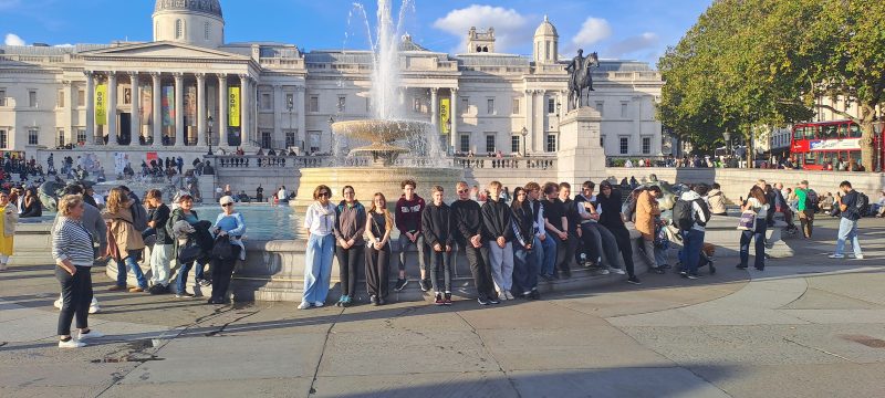 Uczestnicy wycieczki siedzą na obramowaniu fontanny na Trafalgar Squeare. 