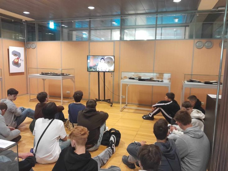 Sala konferencyjna. Uczestnicy siedzą na podłodze i oglądają film na wyświetlany na ekranie telewizora. 