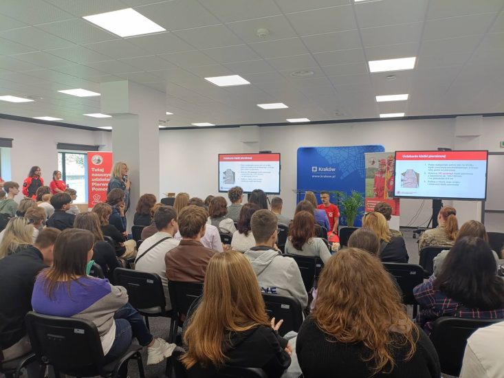 Sala konferencyjna z dwoma ekranami. Uczestnicy (widoczni o tyłu) siedzą i słuchają wykładu .