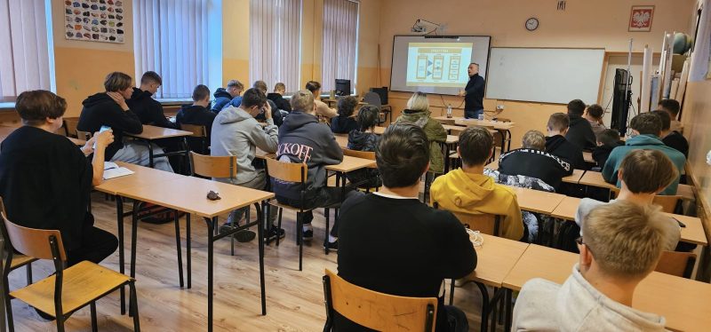 Uczniowie w sali lekcyjnej uczestniczą w zajęciach podczas których prowadzący wyświetla na ekranie prezentację.