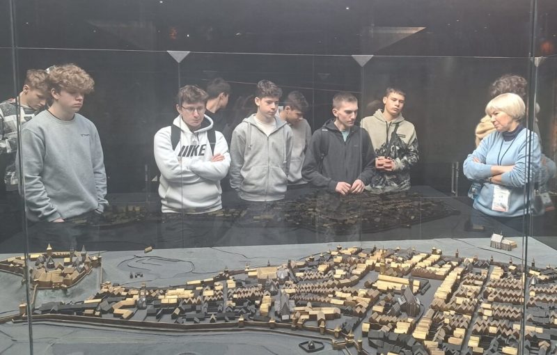 Grupa uczniów stoi przed makieta miasta i słucha wykładu przewodnika.