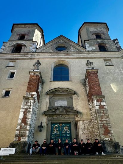 Wejście główne do kościoła w Tyńcu. Widoczna jest cała fasada kościoła z wieżami. Na schodach siedzą uczestnicy wycieczki. 