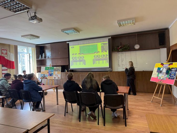 Sala konferencyjna. Uczniowie w grupach siedzą przy stolikach. Na ekranie jest wyświetlana prezentacja. Przy tablica uczennica zapisuje w tabelce punkty.